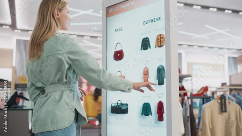 Beautiful Female Customer Using Floor-Standing LCD Touch Display while Shopping in Clothing Store. She is Choosing Stylish Bags, Picking Different Designs from Collection. People in Fashionable Shop. photo