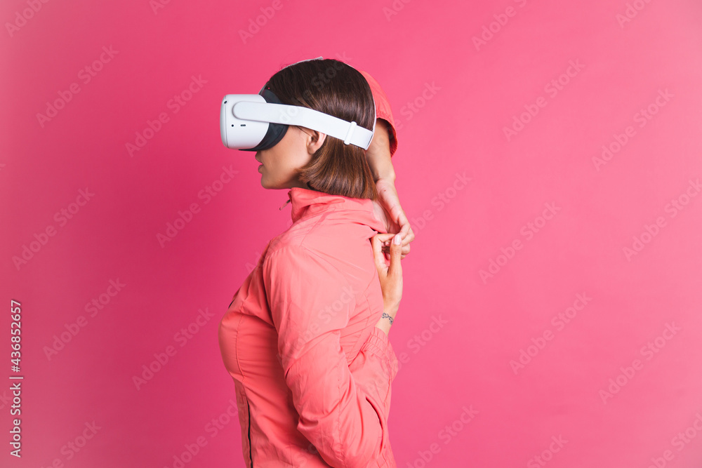 Fototapeta premium Young fit woman in sport wear jacket and virtual reality glasses stretching herself on pink background