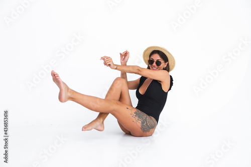 Fit tanned woman in black swimsuit and straw hat on white background posing