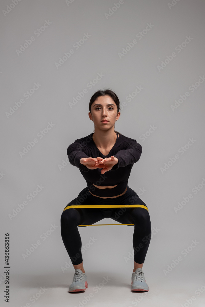 Fit brunette doing squats with resistance band on grey background Fit brunette doing squats with resistance band on grey background