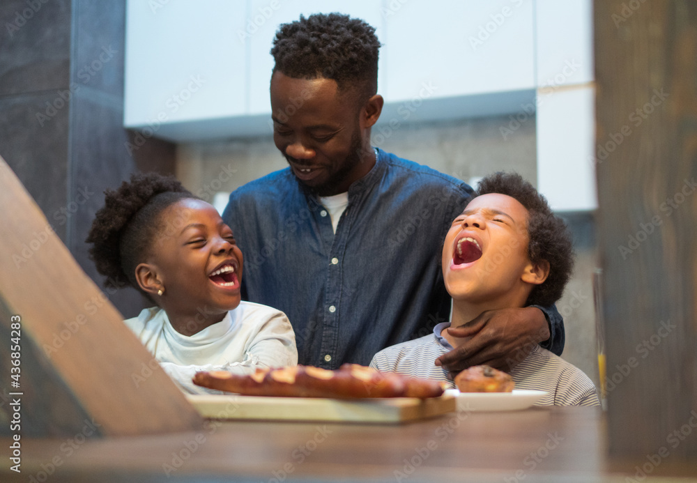 A single father enjoying breakfast time with his two children. They are sitting at the table in the kitchen and having a big lough. - obrazy, fototapety, plakaty 