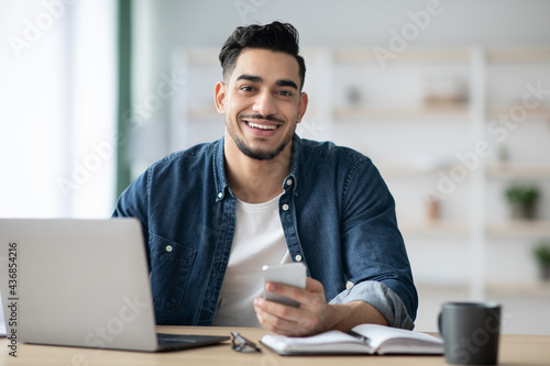 Smiling arab guy using smartphone while sitting at workdesk photo