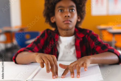 Blind african american schoolboy sitting at desk in classroom reading braille book with fingers