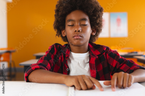 African american schoolboy sitting in classroom with eyes closed reading braille book with fingers