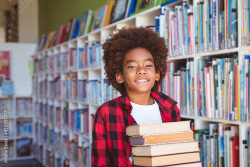 Smiling african american schoolboy carrying stack of books in school library
