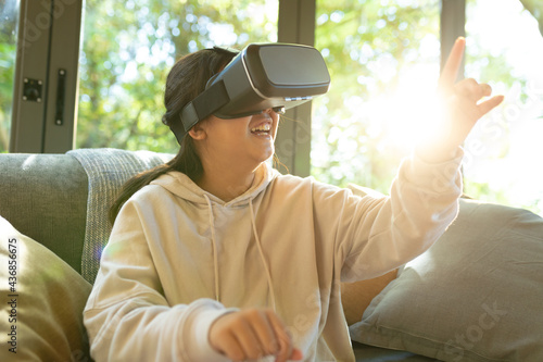 Smiling asian girl wearing vr headset touching with her finger sitting on sofa