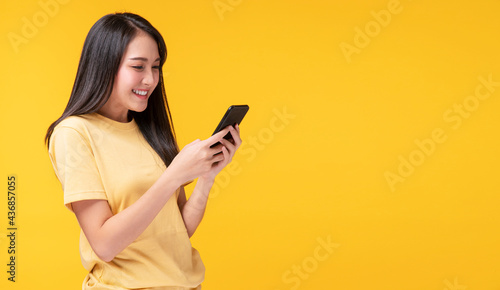 Obraz na plátně Cheerful asian woman holding smartphone and her hand with shocked amazed for success or get good news over isolated yellow background