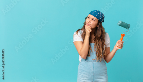 young pretty woman thinking, feeling doubtful and confused and painting a wall