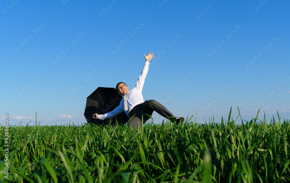 businessman poses with umbrella in a field, he's falling, a concept of crisis, green grass and blue sky as background businessman poses with umbrella in a field, he's falling, a concept of crisis, green grass and blue sky as background
