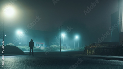 A mysterious hooded man standing in a street on an industrial estate looking at buildings on a misty winters night.