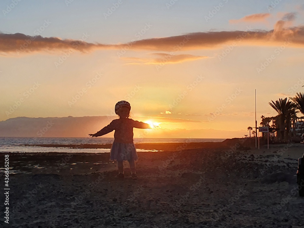 Little toddler girl, standing on the edge of the sea shore on sunset, enjoying the beautiful nature