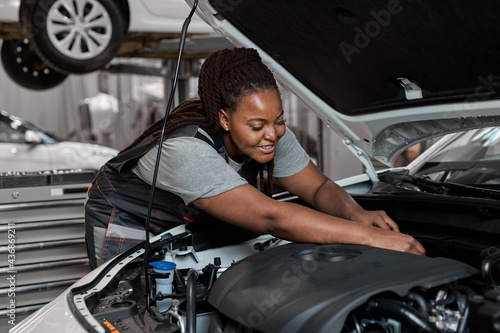 Young african female car repair worker check the oil level in car engine, side view. Smiling black mechanic checking and maintenance car engine or vehicle with herrself. Copy space © Roman
