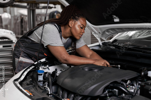 african woman under the hood of white car. woman in uniform mechanic repairing a car in car service. portrait of young black female enjoying work with automobile, vehicle