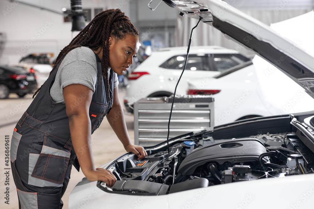 Afro Car mechanic woman is examining under hood of car at repair garage,  wearing overalls, looking confident and concentrated. Side view on female  trying to solve the problem of inoperative engine Photos