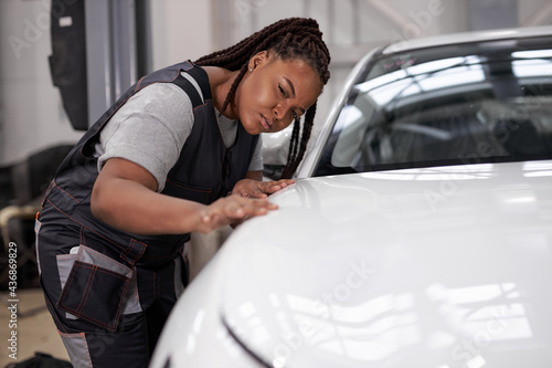 Confident Afro american Female auto mechanic touching surface of white repainted and clean car body in auto repair shop. Pretty black woman Mechanic in uniform working in workshop, looking serious
