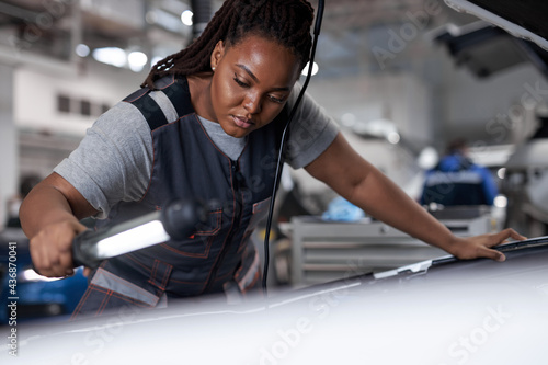 black woman mechanic looking to car engine and holding lamp, Car master in service shop, female mechanic repairer service technician checks and repairs the engine condition under hood of vehicle