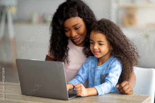 Home schooling during covid concept. Cheerful black mom helping her daughter with online homework on laptop pc