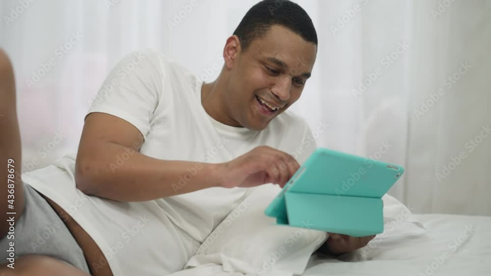 Young African American male millennial surfing social media on tablet laughing lying on bed at home. Portrait of positive joyful handsome man resting in bedroom on weekend morning indoors Young African American male millennial surfing social media on tablet laughing lying on bed at home. Portrait of positive joyful handsome man resting in bedroom on weekend morning indoors