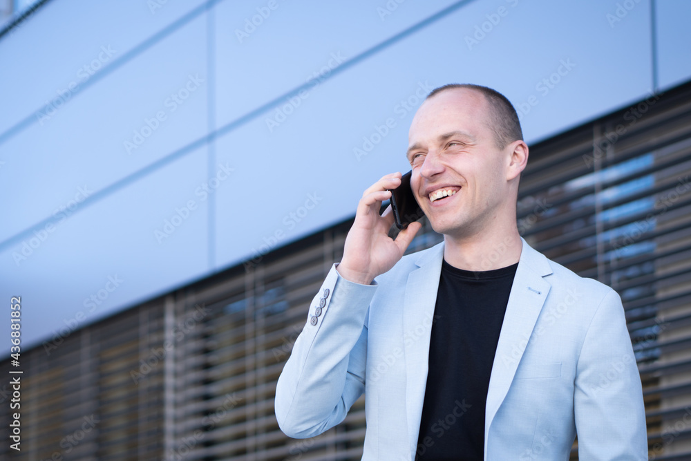Bottom view perspective of young short-haired businessman in a blue suit standing in front of a corporate building solves a business phone call. Bottom view perspective of young short-haired businessman in a blue suit standing in front of a corporate building solves a business phone call.