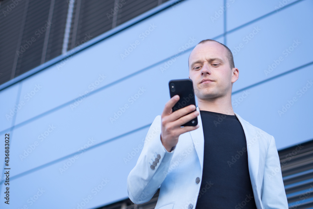 Bottom view perspective of young short-haired businessman in a blue suit standing in front of a corporate building and writes a text message on mobile. Bottom view perspective of young short-haired businessman in a blue suit standing in front of a corporate building and writes a text message on mobile.
