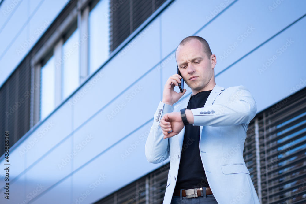 Bottom view perspective of young short-haired businessman in a blue suit standing in front of a corporate building solves a business phone call. Bottom view perspective of young short-haired businessman in a blue suit standing in front of a corporate building solves a business phone call.