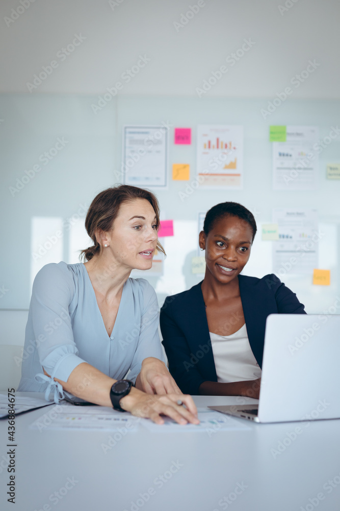Two diverse businesswomen sitting at desk, using laptop, talking Two diverse businesswomen sitting at desk, using laptop, talking