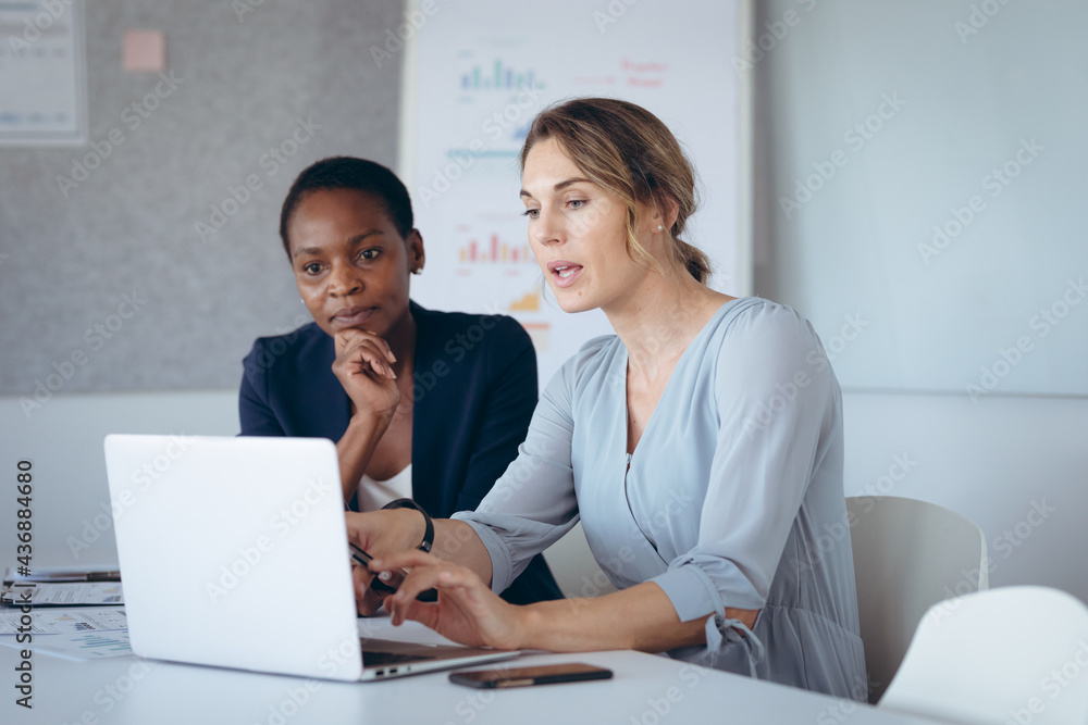 Two diverse businesswomen sitting at desk, using laptop, talking Two diverse businesswomen sitting at desk, using laptop, talking