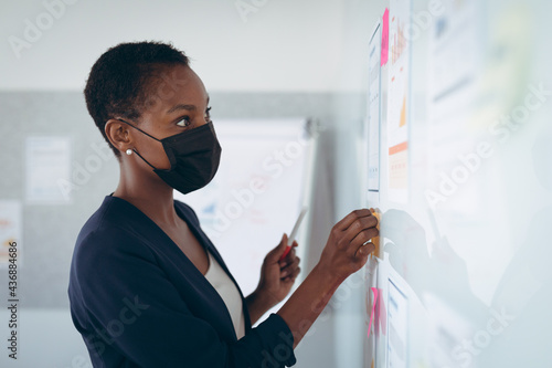 African american businesswoman wearing face mask making notes and adding post-ins on wall photo