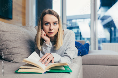Young beautiful blonde woman reads an interesting book while lying on the couch, resting after work