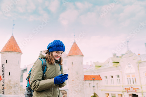 Tallinn, Estonia. Young Caucasian Woman Backpacker Tourist Enjoying Life, Smiling And Using Smartphone Near Landmark Viru Gate. Vacation In Old Town. Girl Surfing Net Internet In Phone