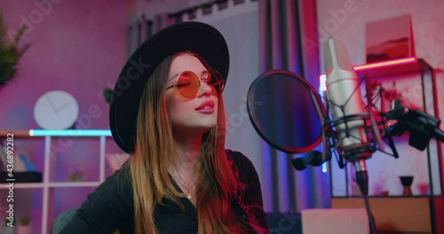 Charming creative successful young female singer with long straight hair in stylish hat and sunglasses singing song in front of recording equippment to the sounds of her guitar in home studio on the photo