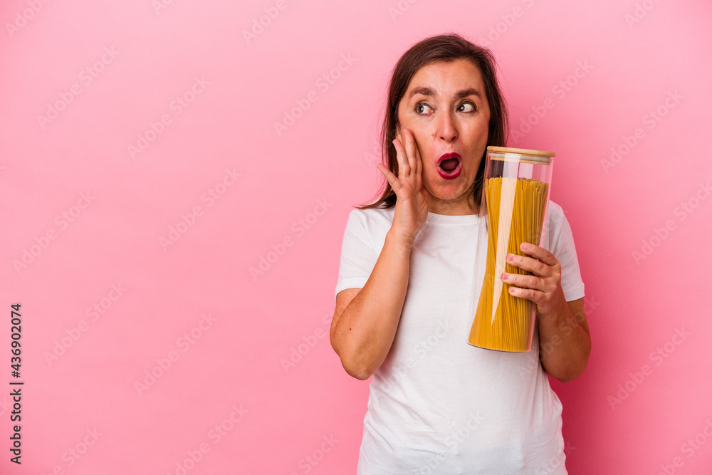 Middle age caucasian woman holding a pasta jar isolated on pink background is saying a secret hot braking news and looking aside Middle age caucasian woman holding a pasta jar isolated on pink background is saying a secret hot braking news and looking aside
