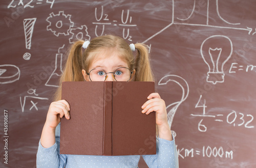 Little girl hiding behind the book standing near chalkboard full of signs and formulas. Childhood and education concept.