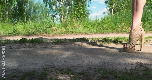 woman confidently walks in high-heeled sandals along an earthen path in nature along green trees, clear day, bright sun, close-up of legs, slow motion
