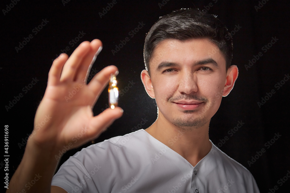 Obraz premium A young man who is a medical doctor in a white uniform poses against a black background in the studio with an ampoule in his hand