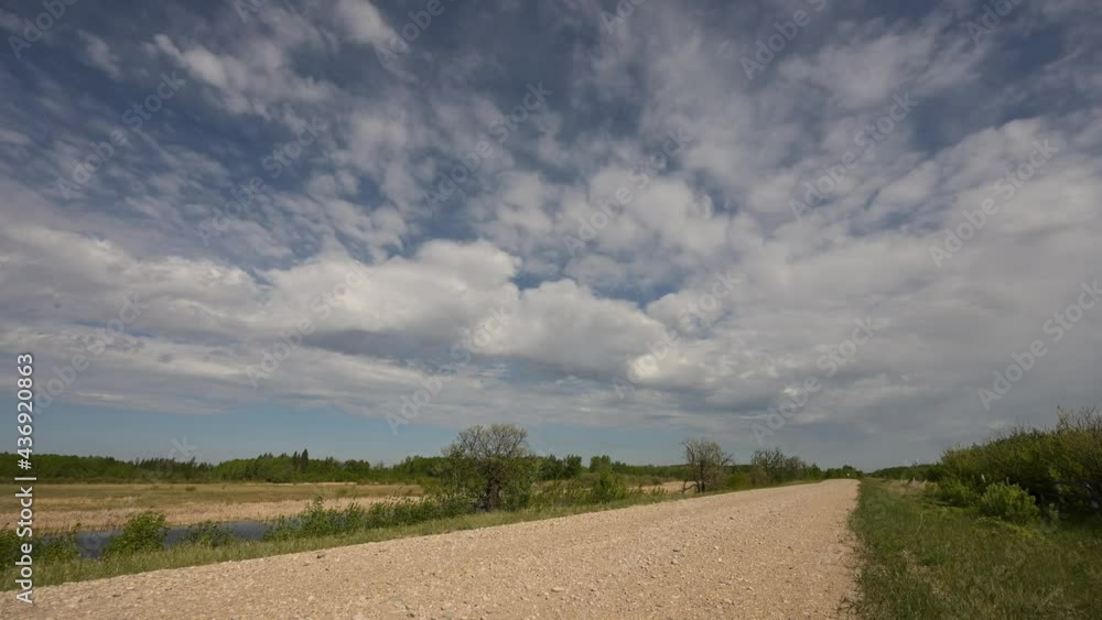 Time lapse of white clouds in a blue sky moving towards view over a gravel farm road.
Time lapse of white clouds in a blue sky moving towards view over a gravel farm road.