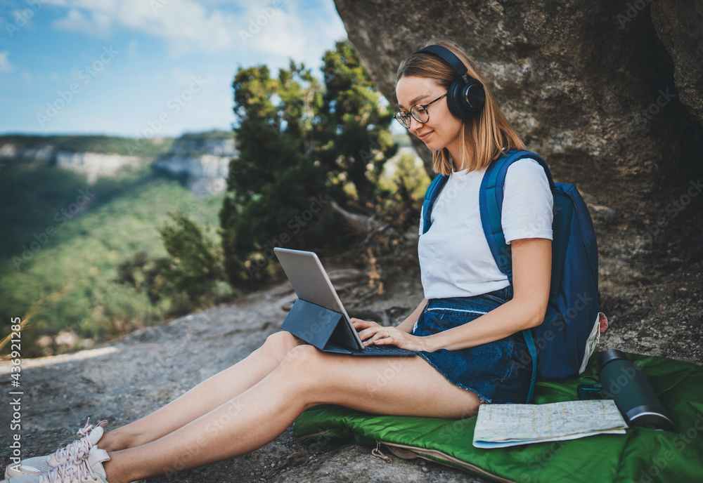 cute hiker girl sitting in mountains and listening to music with headphones using laptop in nature, traveler girl with backpack looks at map for planning route using online Internet in tablet outside