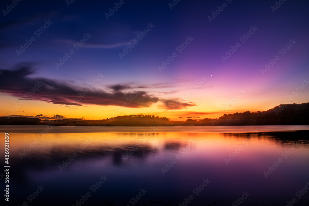 Uma paisagem focada no pôr-do-sol com montanhas e um lago espelhado e cristalino com cores vibrantes azul, amarelo, magenta e laranja com algumas nuvens contrastantes com a paisagem.