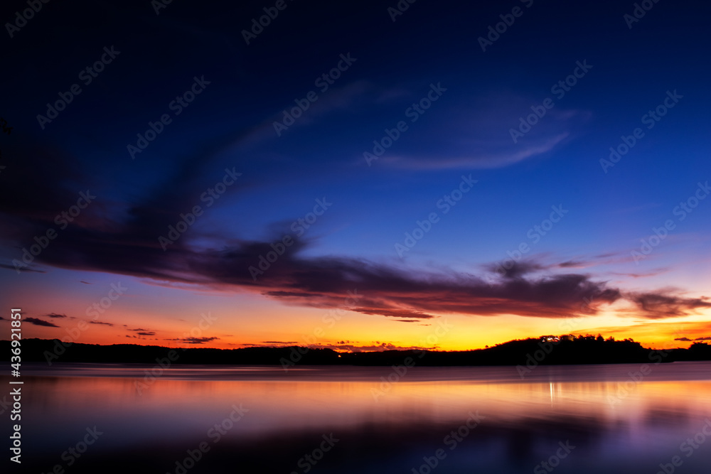 Uma paisagem focada no pôr-do-sol com montanhas e um lago espelhado e cristalino com cores vibrantes azul, amarelo, magenta e laranja com algumas nuvens contrastantes com a paisagem.