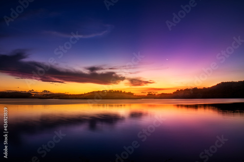 Uma paisagem focada no pôr-do-sol com montanhas e um lago espelhado e cristalino com cores vibrantes azul, amarelo, magenta e laranja com algumas nuvens contrastantes com a paisagem.