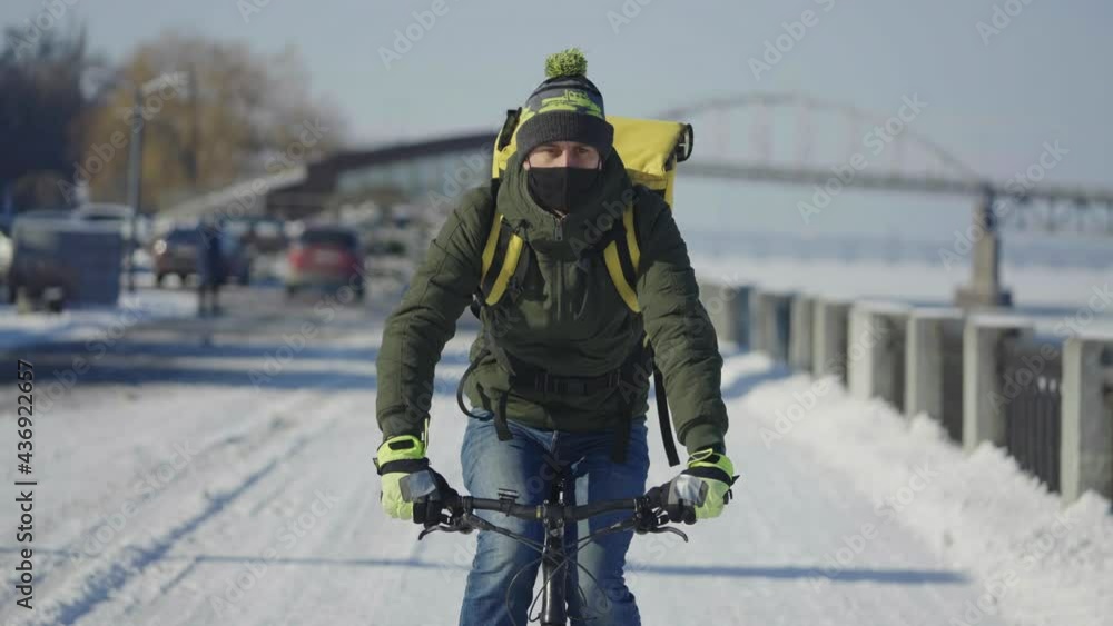 Portrait of cyclist with backpack and protective mask rides bicycle on snow covered street with food delivery. Man works as courier and delivers order to customers during virus outbreak. Slow motion. Portrait of cyclist with backpack and protective mask rides bicycle on snow covered street with food delivery. Man works as courier and delivers order to customers during virus outbreak. Slow motion.