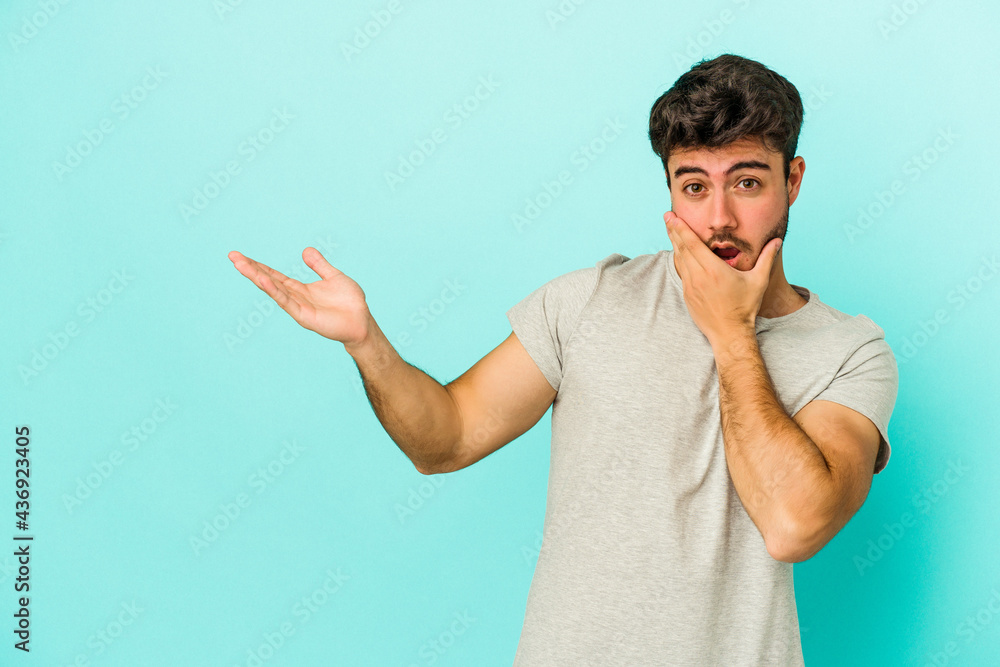 Young caucasian man isolated on blue background holds copy space on a palm, keep hand over cheek. Amazed and delighted. Young caucasian man isolated on blue background holds copy space on a palm, keep hand over cheek. Amazed and delighted.
