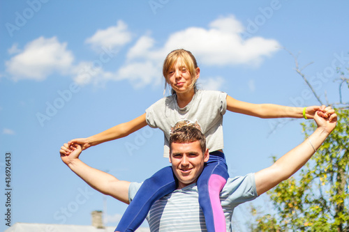 Defocused brother riding sister on back. Portrait of happy girl on man shoulders, piggyback. Girl fly, raise hands. Family playing outside. Blue green nature background. Summer mood. Out of focus