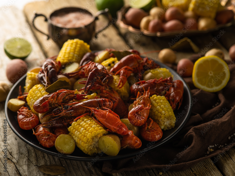 In the foreground of the photo, we see a dish with boiled corn and crayfish. In the background is a photo of a gravy boat with sauce, lemon and vegetables. Wooden table. Country style. In the foreground of the photo, we see a dish with boiled corn and crayfish. In the background is a photo of a gravy boat with sauce, lemon and vegetables. Wooden table. Country style.