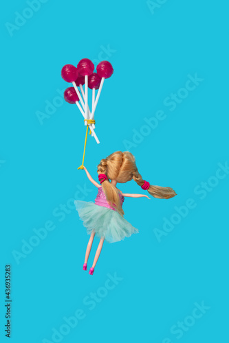 Child doll holding purple red lollipops as balloons flying against sky blue background. Minimal creative composition.