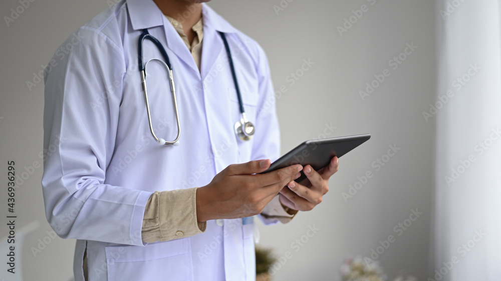 Cropped shot of A medical professional in a lab gown with a stethoscope holding a tablet in hand, side view shot. Cropped shot of A medical professional in a lab gown with a stethoscope holding a tablet in hand, side view shot.