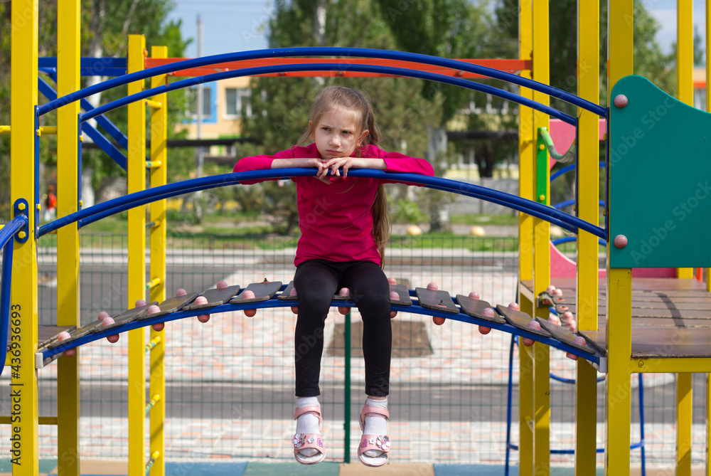 Fototapeta premium A child girl with blond hair in a pink T-shirt is sad on the playground, sits on a slide, her legs dangling. 
