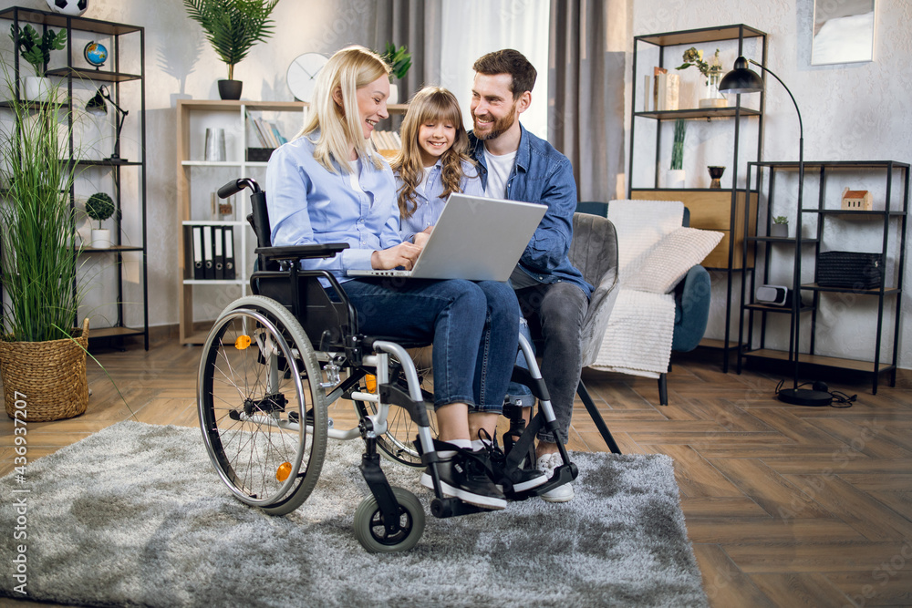 Pretty little kid with father and disabled mother using modern laptop during free time at home. Female blonde sitting in wheelchair. Casual day of young family. Pretty little kid with father and disabled mother using modern laptop during free time at home. Female blonde sitting in wheelchair. Casual day of young family.
