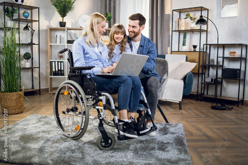 Pretty little kid with father and disabled mother using modern laptop during free time at home. Female blonde sitting in wheelchair. Casual day of young family.