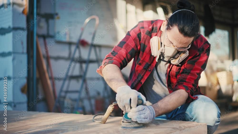 A young attractive man in a construction respirator grinds a parquet board with an orbital sander outdoors. A young attractive man in a construction respirator grinds a parquet board with an orbital sander outdoors.